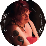 Person with short red hair and a black tattoo of a stylized semicolon and planet on their upper arm, smiling and holding a pink object, against a dark background. Wearing a black top and a necklace.